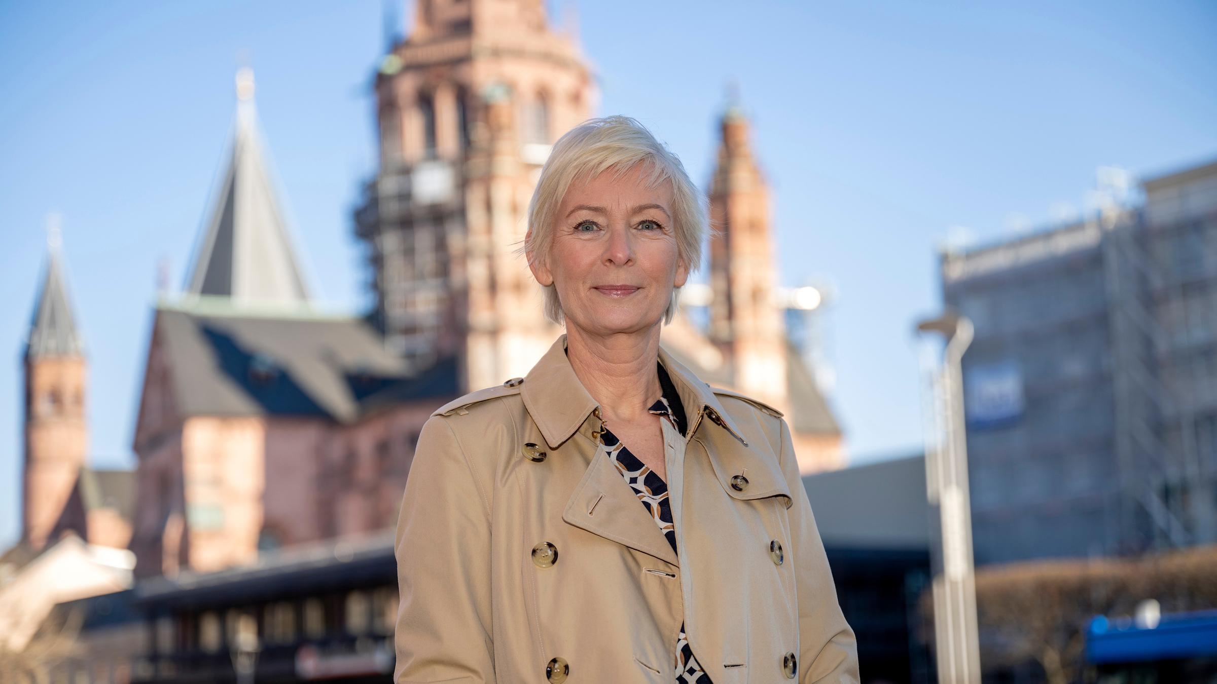 Die Mainzer Stadtschreiberin Dörte Hansen steht im Freien und trägt einen beigen Trenchcoat. Im Hintergrund ist der Mainzer Dom zu erkennen. 