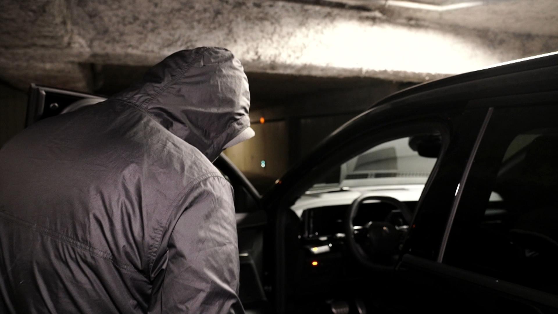 Ein Mann mit Kapuze steht vor einem Auto und versucht, das Auto zu knacken. 