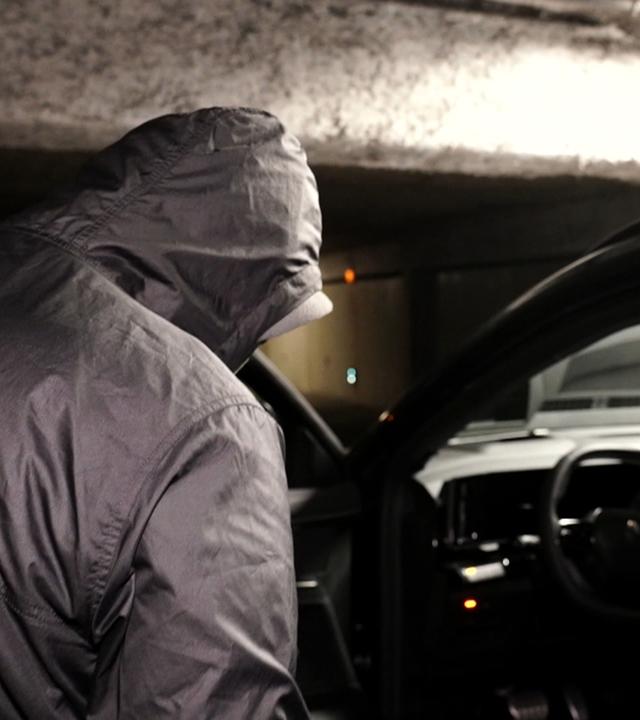 Ein Mann mit Kapuze steht vor einem Auto und versucht, das Auto zu knacken. 