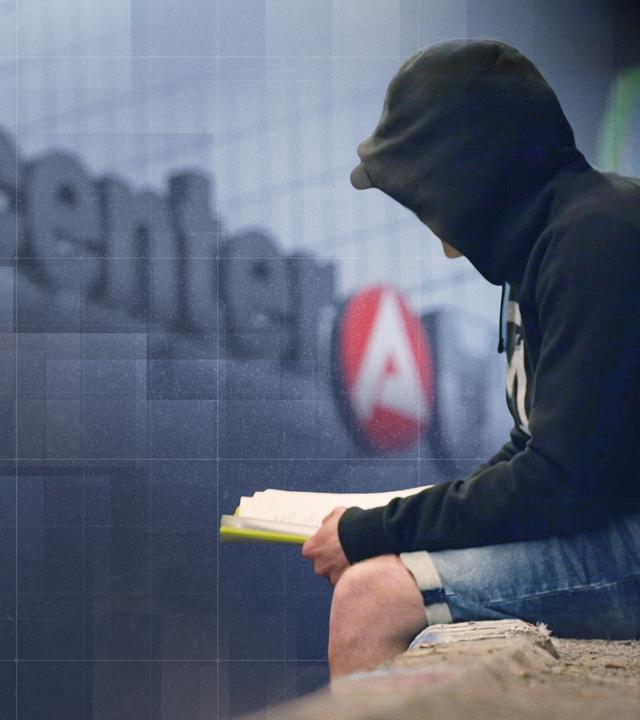Mann mit schwarzem Hoodie sitzt vor einem Gebäude mit "jobcenter"-Schriftzug