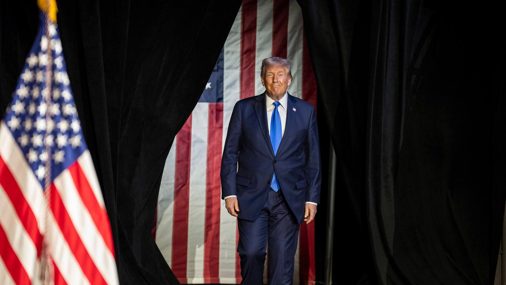 Ein älterer Mann im blauen Anzug tritt durch einen schwarzen Vorhand. Im Hintergrund sind Flaggen der USA zu sehen.