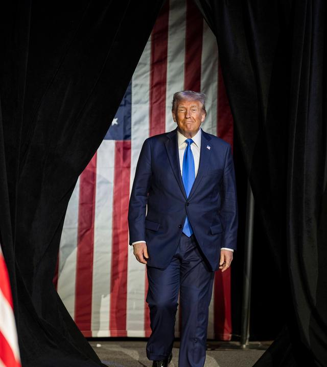 Ein älterer Mann im blauen Anzug tritt durch einen schwarzen Vorhand. Im Hintergrund sind Flaggen der USA zu sehen.