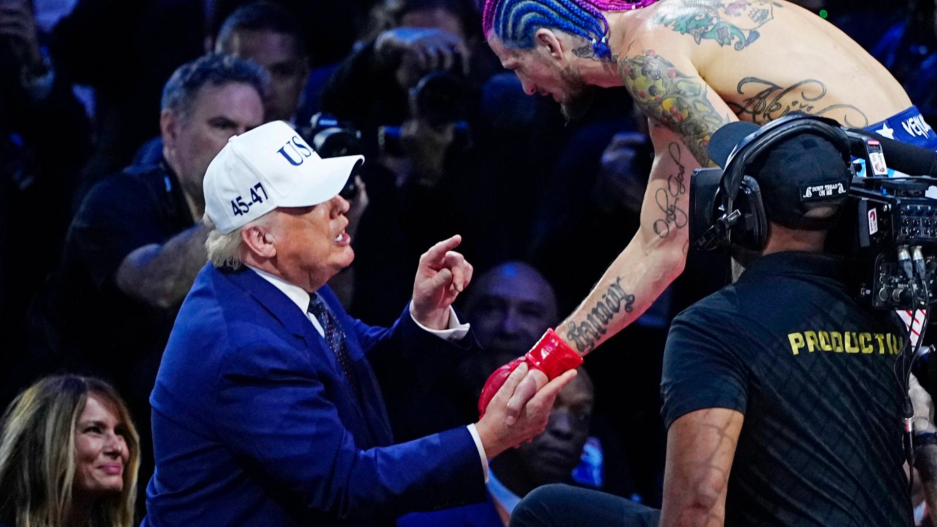 US-Präsident Donald Trump, ein älterer Mann mit Baseball-Kappe und im dunkelblauen Anzug gibt einem MMA-Kämpfer mit nacktem tätowierten Oberkörper und gefärbten Haaren die Hand, während er mit dem Finger auf ihn zeigt und redet.