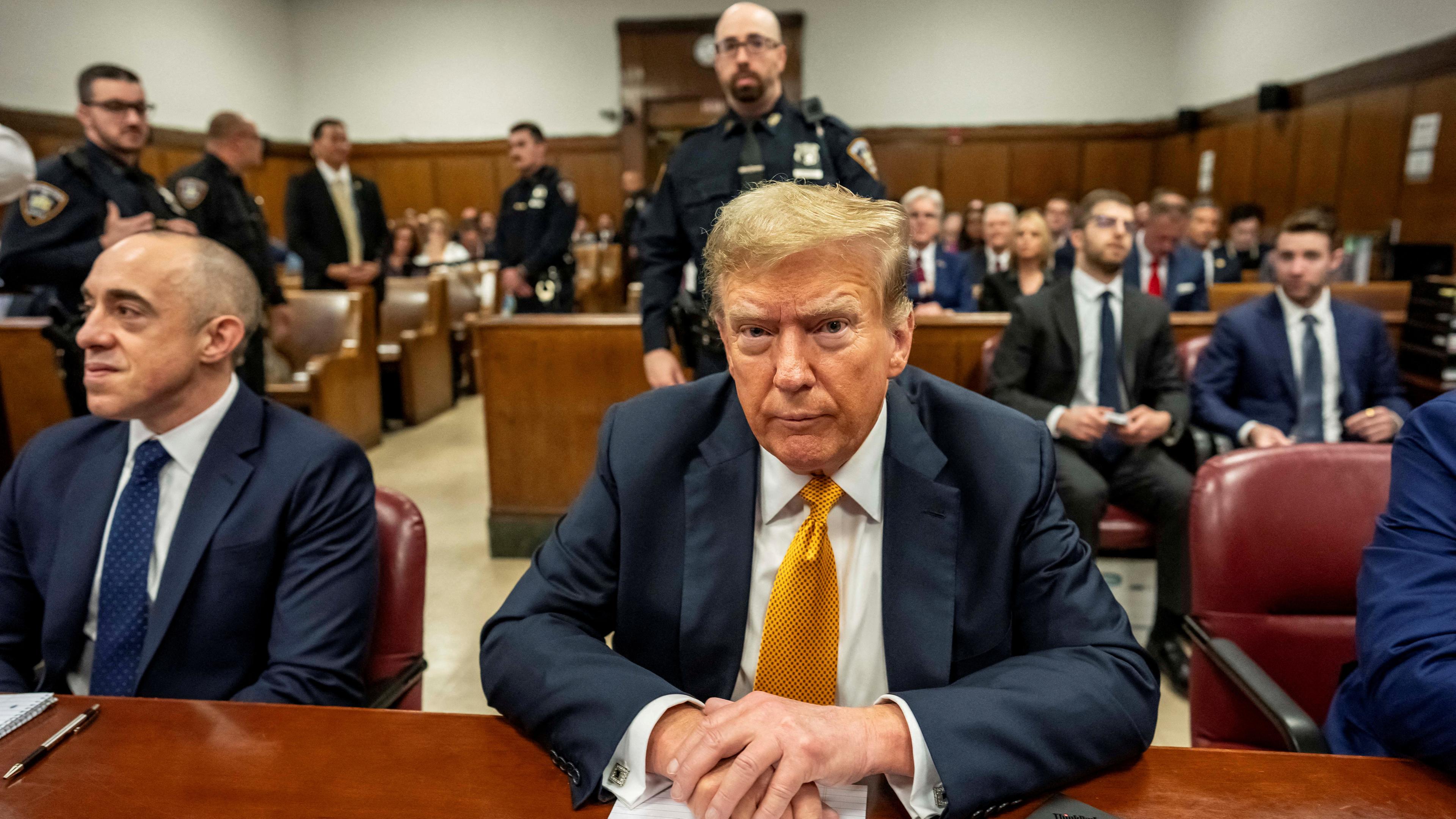 Der ehemalige US-Präsident Donald Trump sitzt in einem blauen Anzug mit goldener Kravatte auf der Anklagebank.