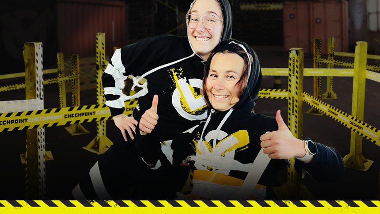 Checkpoint: Die Testteamerinnen Laura und Maja stehen vor einem abgesteckten Parcours und sind mit doppelseitigem Klebeband eingewickelt. Beide schauen fröhlich in die Kamera. 
