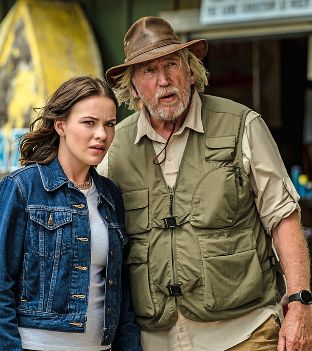 Issie (rechts) und Gordon (links) stehen vor einem Bootsverleih und schauen mit ernstem Blick in Richtung des Strandes.