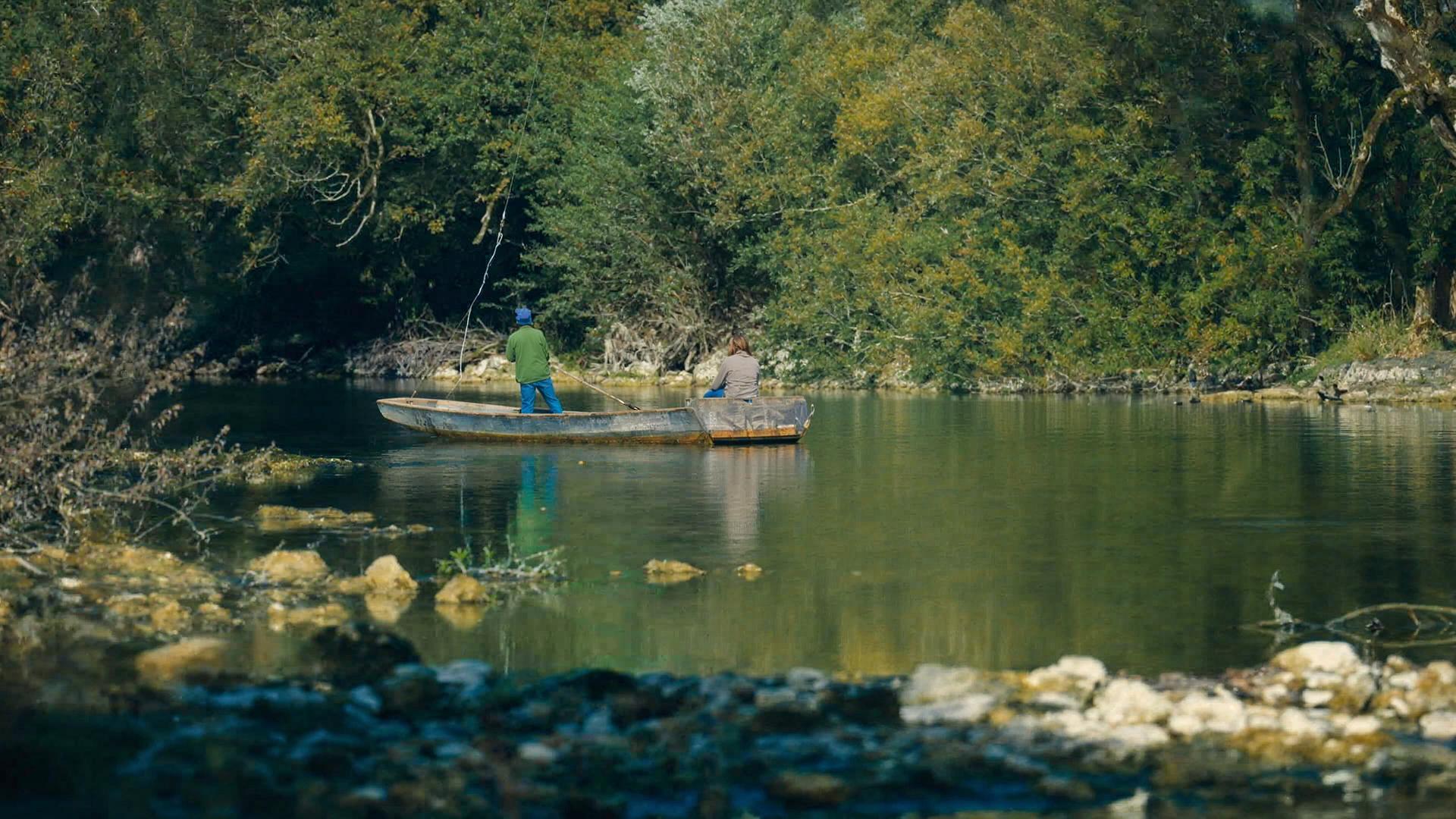 Zwei Personen angeln in einem ruhigen Abschnitt des Grenzflusses Doubs, umgeben von üppiger Vegetation. Sie befinden sich in einem flachen Boot, das sanft auf dem klaren Wasser treibt, während die Natur in herbstlichen Farben leuchtet.