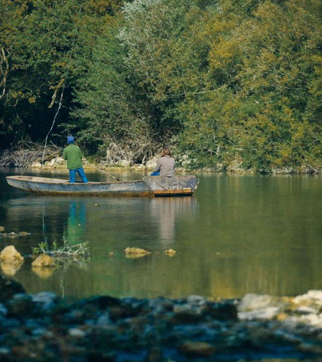 Zwei Personen angeln in einem ruhigen Abschnitt des Grenzflusses Doubs, umgeben von üppiger Vegetation. Sie befinden sich in einem flachen Boot, das sanft auf dem klaren Wasser treibt, während die Natur in herbstlichen Farben leuchtet.