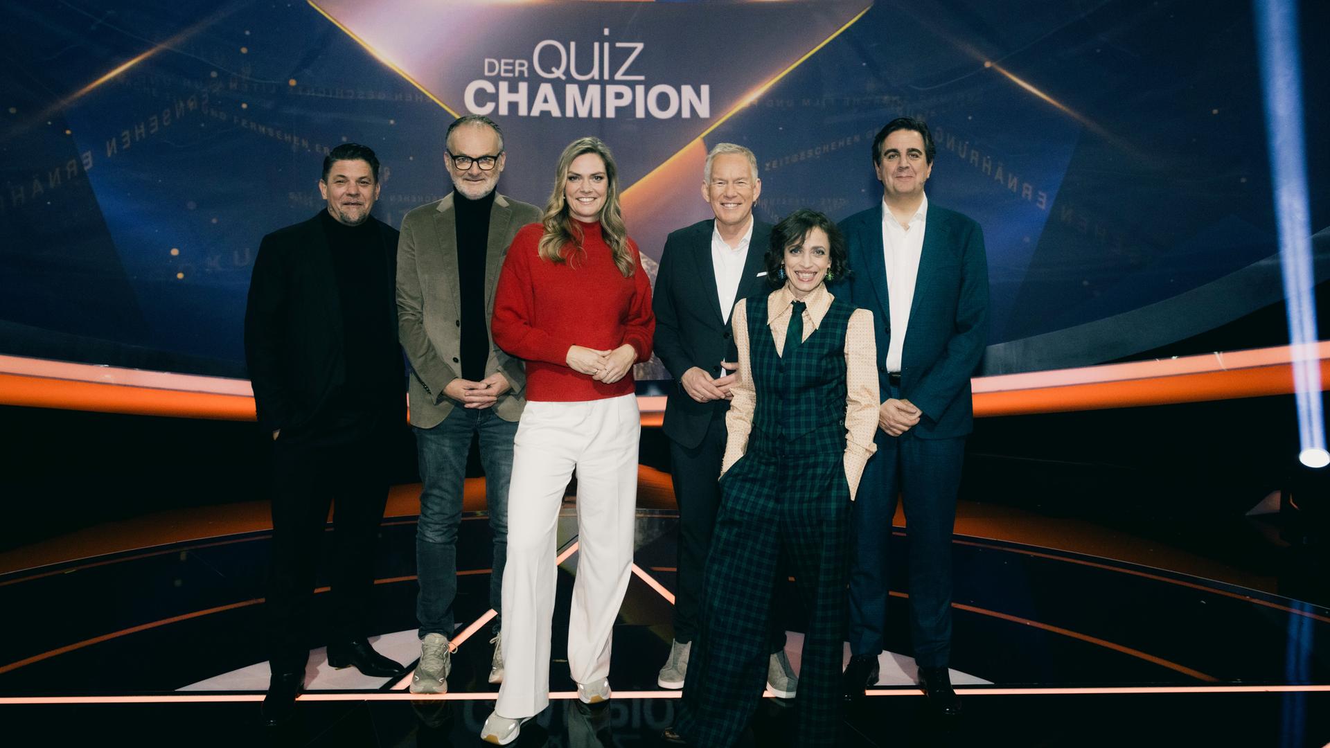 "Der Quiz-Champion": Gruppenfoto: Tim Mälzer, Frank Plasberg, Julia Scharf, Johannes B. Kerner, Anna Thalbach, Bastian Pastewka stehen vor dem Sendungslogo im Studio und lächeln in die Kamera.