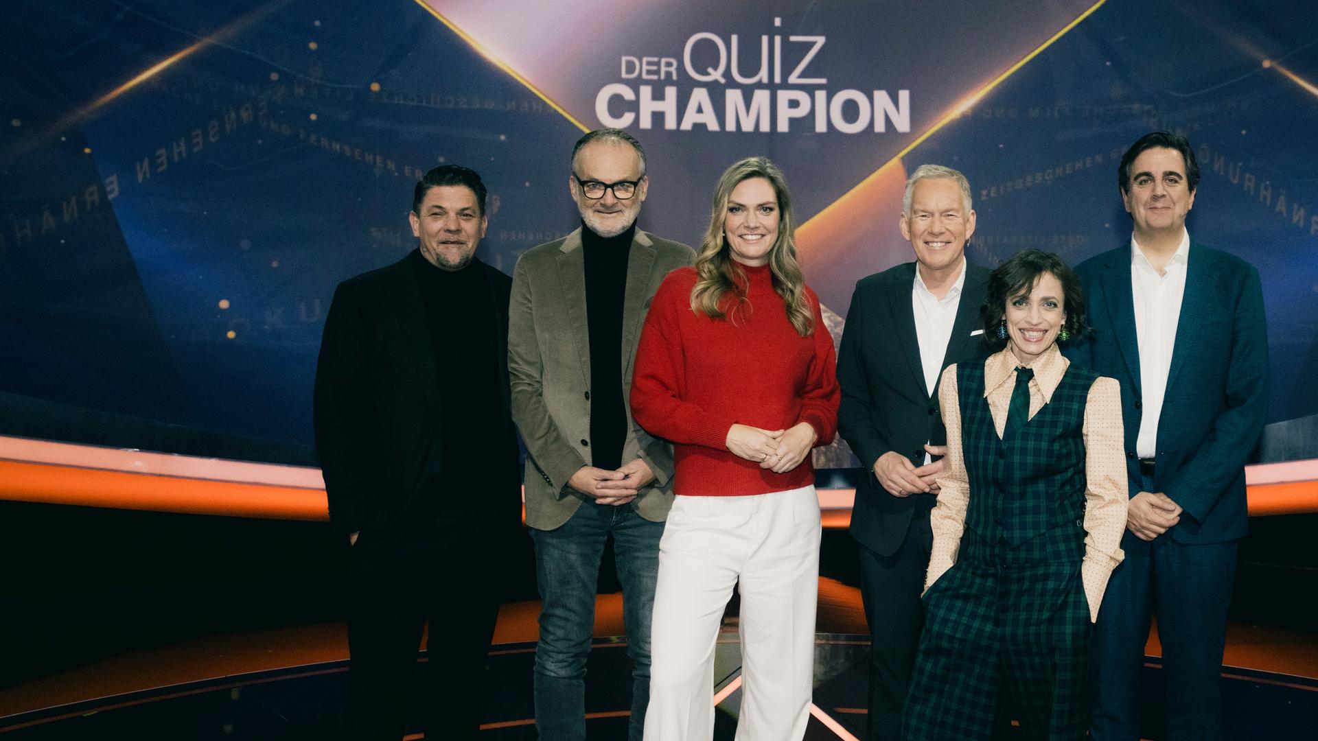 "Der Quiz-Champion": Gruppenfoto: Tim Mälzer, Frank Plasberg, Julia Scharf, Johannes B. Kerner, Anna Thalbach und Bastian Pastewka stehen vor dem Sendungslogo im Studio und lächeln in die Kamera.