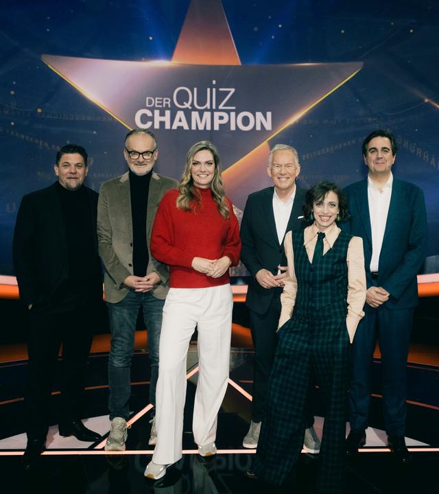 "Der Quiz-Champion": Gruppenfoto: Tim Mälzer, Frank Plasberg, Julia Scharf, Johannes B. Kerner, Anna Thalbach, Bastian Pastewka stehen vor dem Sendungslogo im Studio und lächeln in die Kamera.