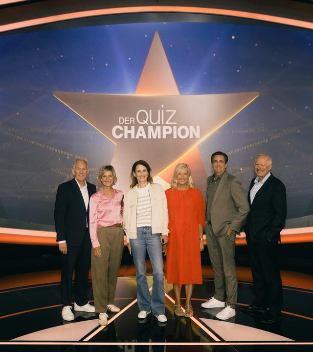 "Der Quiz-Champion": Johannes B. Kerner, Barbara Hahlweg, Katrin Müller-Hohenstein, Andrea Kiewel, Bastian Pastewka und Axel Milberg stehen vor dem Sendungslogo auf der Bühne und lächeln in die Kamera.