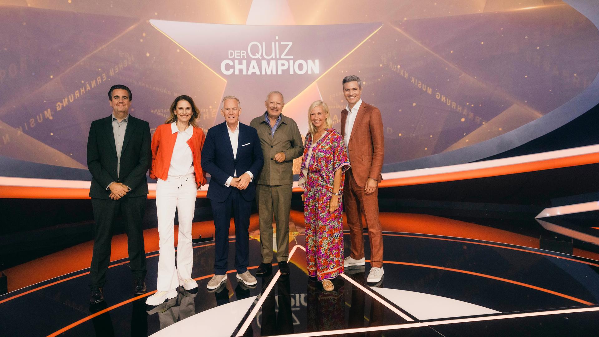 "Der Quiz-Champion": Bastian Pastewka, Katrin Müller-Hohenstein, Johannes B. Kerner, Axel Milberg, Andrea Kiewel und Ingo Zamperoni stehen auf der Bühne vor dem Sendungslogo und lächeln in die Kamera.