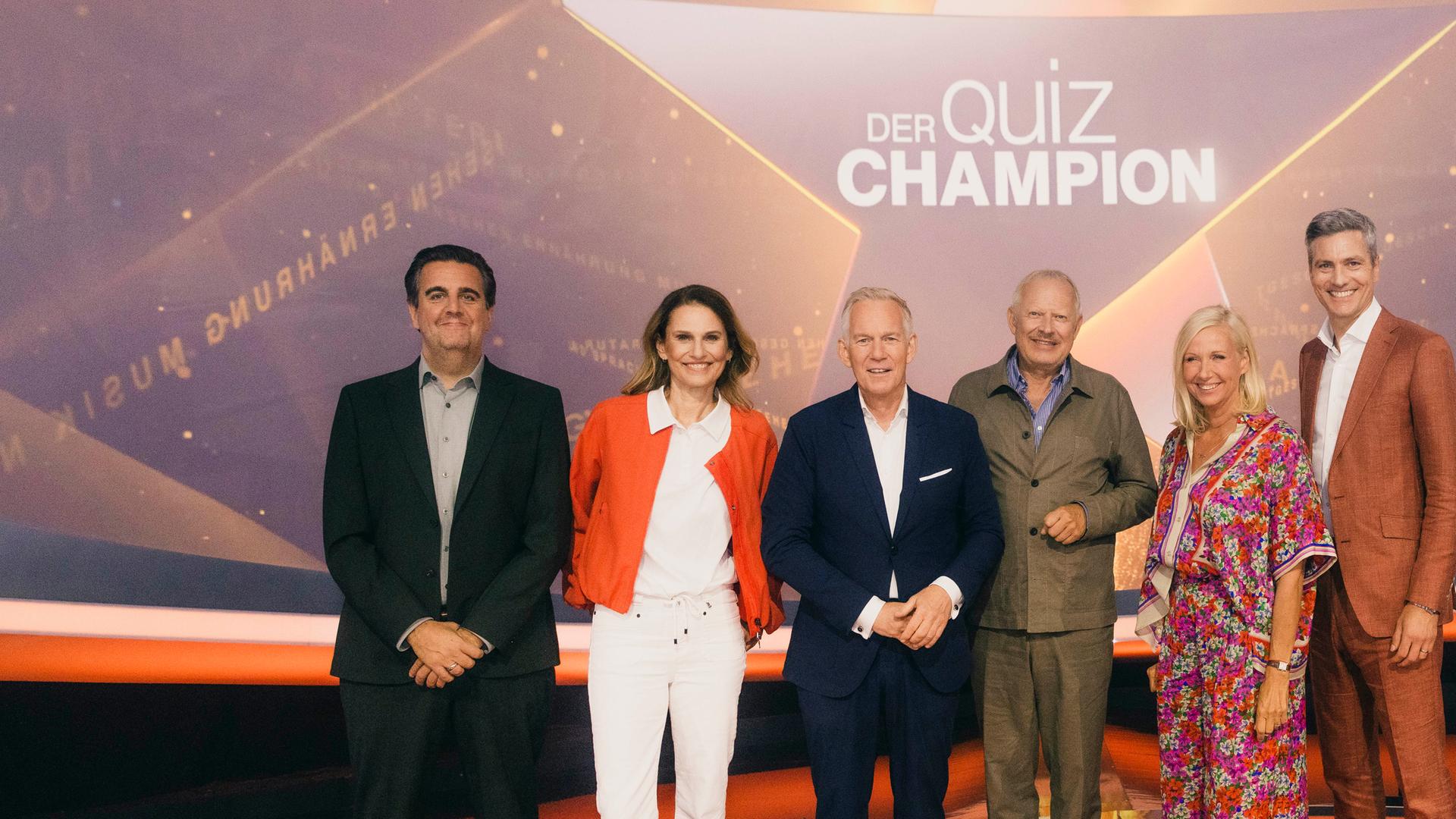 "Der Quiz-Champion": Bastian Pastewka, Katrin Müller-Hohenstein, Johannes B. Kerner, Axel Milberg, Andrea Kiewel und Ingo Zamperoni stehen auf der Bühne vor dem Sendungslogo und lächeln in die Kamera.