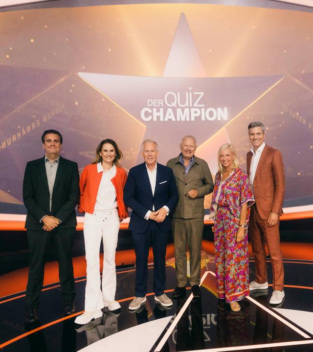 "Der Quiz-Champion": Bastian Pastewka, Katrin Müller-Hohenstein, Johannes B. Kerner, Axel Milberg, Andrea Kiewel und Ingo Zamperoni stehen auf der Bühne vor dem Sendungslogo und lächeln in die Kamera.
