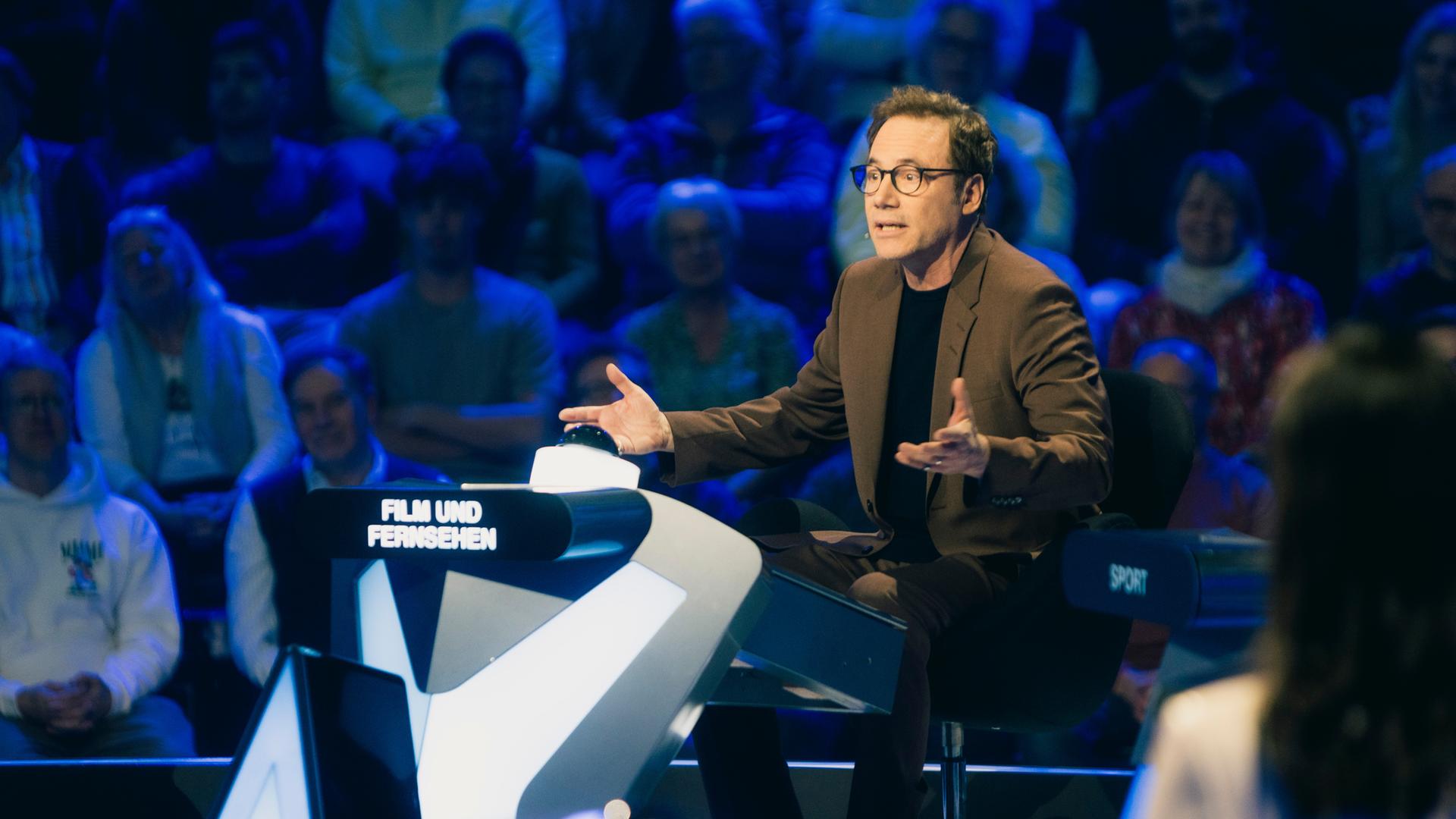 "Der Quiz-Champion": Michael "Bully" Herbig sitzt im Studio an einem Pult und gestikuliert, hinter ihm befindet sich das Publikum.
