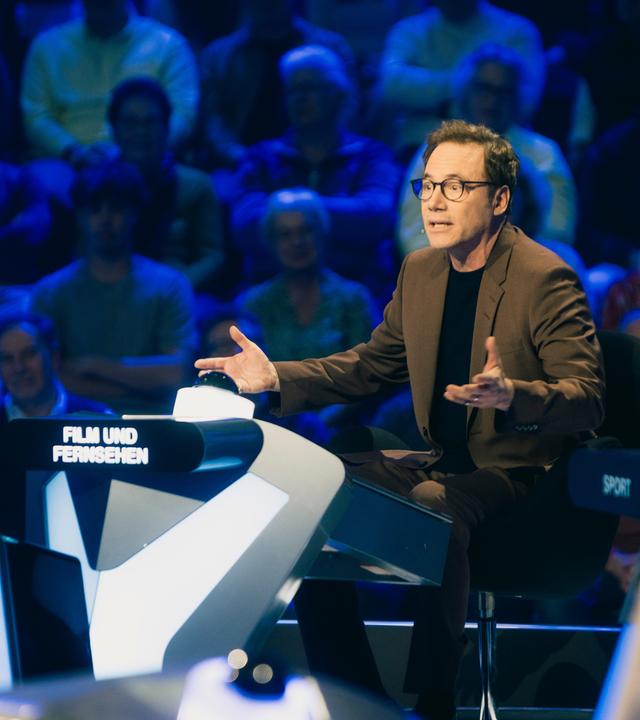"Der Quiz-Champion": Michael "Bully" Herbig sitzt im Studio an einem Pult und gestikuliert, hinter ihm befindet sich das Publikum.
