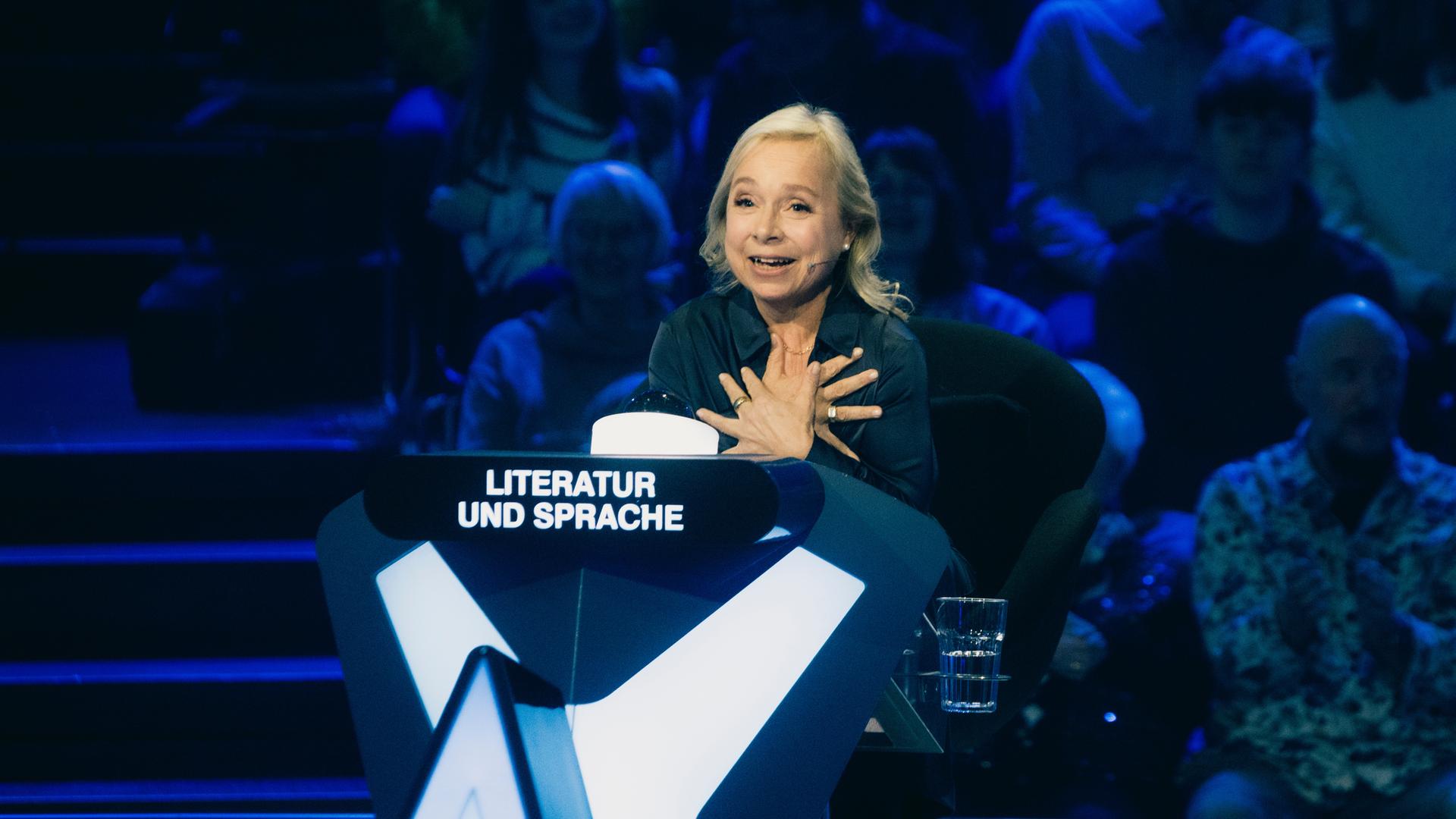 "Der Quiz-Champion": ChrisTine Urspruch sitzt am Ratepult im Studio und gestikuliert.
