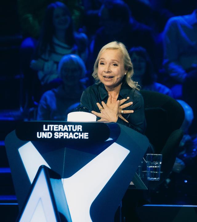 "Der Quiz-Champion": ChrisTine Urspruch sitzt am Ratepult im Studio und gestikuliert.