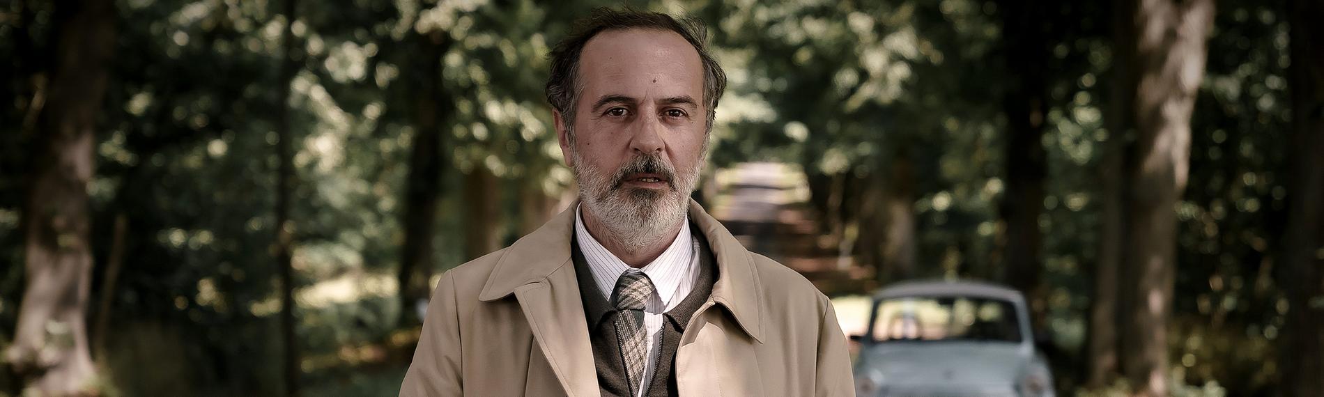 Ballouz (Merab Ninidze) steht auf einer Allee, hat die Hände in der Tasche seines Mantels und guckt nach vorne zum Wegesrand.