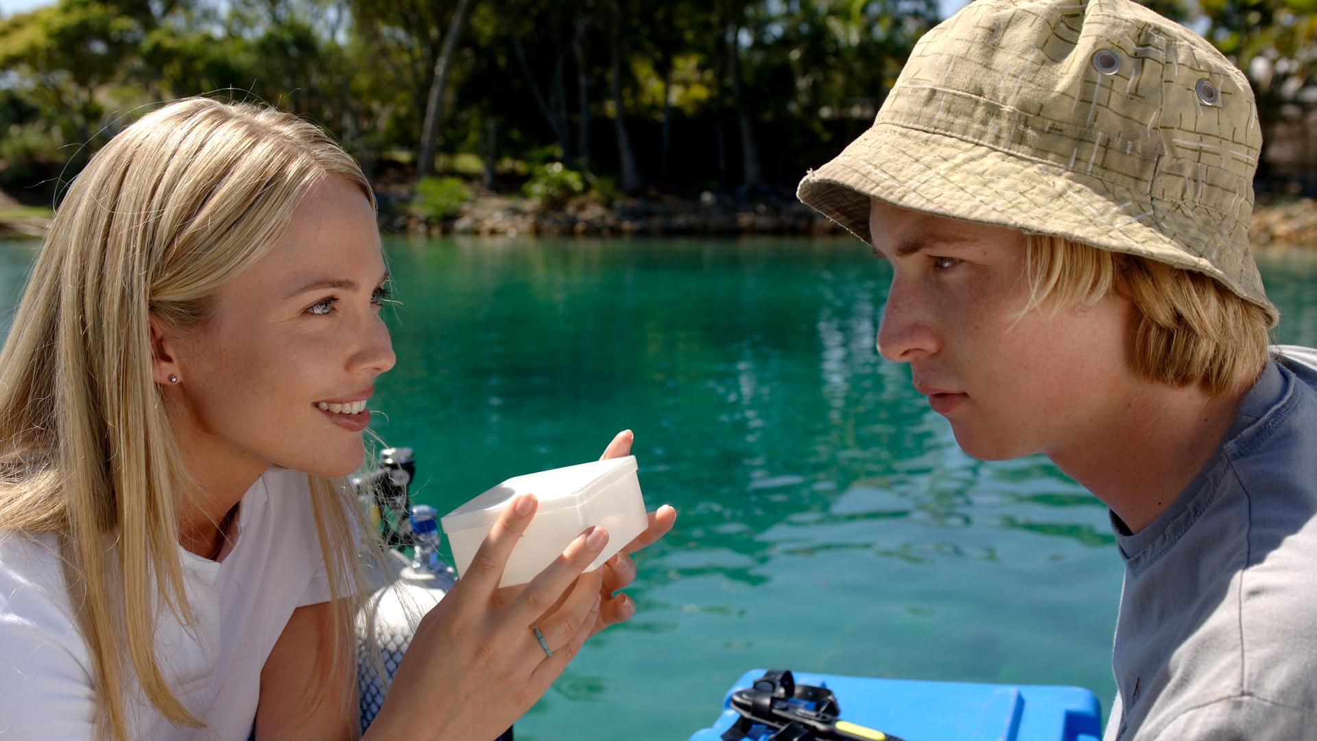 Lewis unterhält sich mit der Meeresbiologin. Lewis (rechts) trägt einen beigefarbenen Fischerhut und schaut die Meeresbiologin (links) an, die eine weiße kleine Schachtel in der Hand hält und ihn anlächelt.