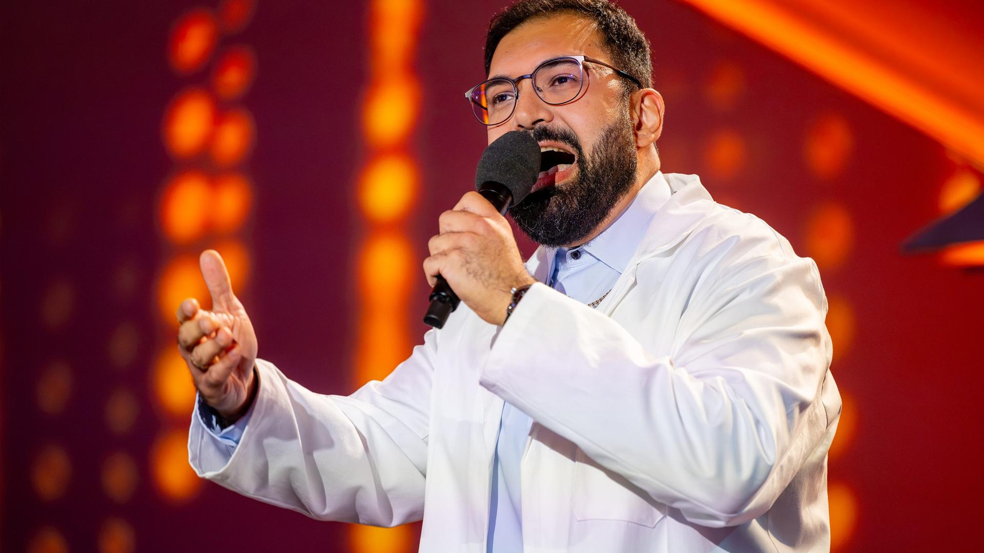 Dr. Emir trägt einen weißen Kittel und ein blaues Hemd darunter. In seiner linken Hand hält er das Mikrofon. Der Hintergrund ist rot beleuchtet. Dr. Emir trägt eine Brille.