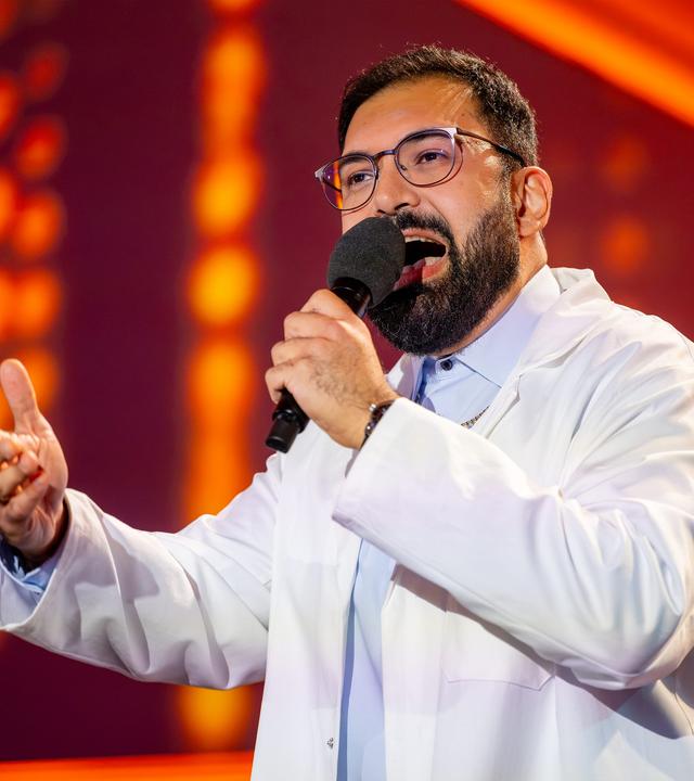 Dr. Emir trägt einen weißen Kittel und ein blaues Hemd darunter. In seiner linken Hand hält er das Mikrofon. Der Hintergrund ist rot beleuchtet. Dr. Emir trägt eine Brille.