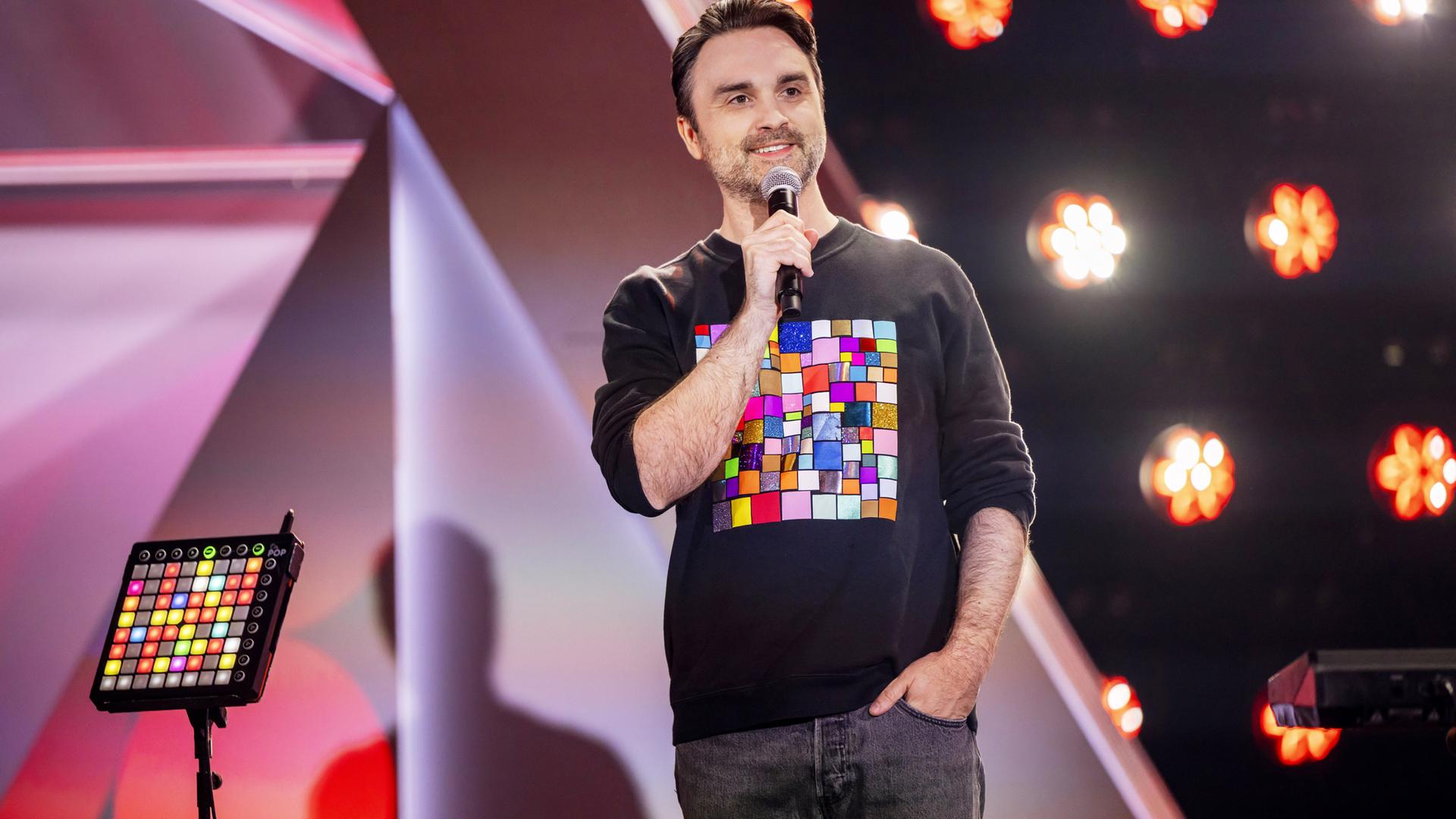 Dr. Pop steht in der Mitte des Bildes. Er trägt einen schwarzen Pullover mit bunten Vierecken darauf. In seiner rechten Hand hält er ein Mikrofon. Seine linke Hand hat er in die Hosentasche gesteckt. Im Hintergrund sind Scheinwerfer zu sehen.