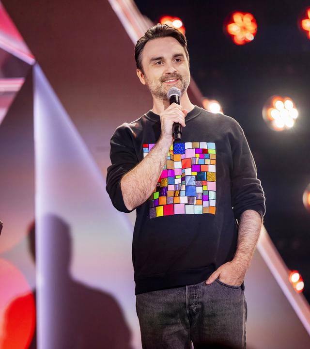 Dr. Pop steht in der Mitte des Bildes. Er trägt einen schwarzen Pullover mit bunten Vierecken darauf. In seiner rechten Hand hält er ein Mikrofon. Seine linke Hand hat er in die Hosentasche gesteckt. Im Hintergrund sind Scheinwerfer zu sehen.