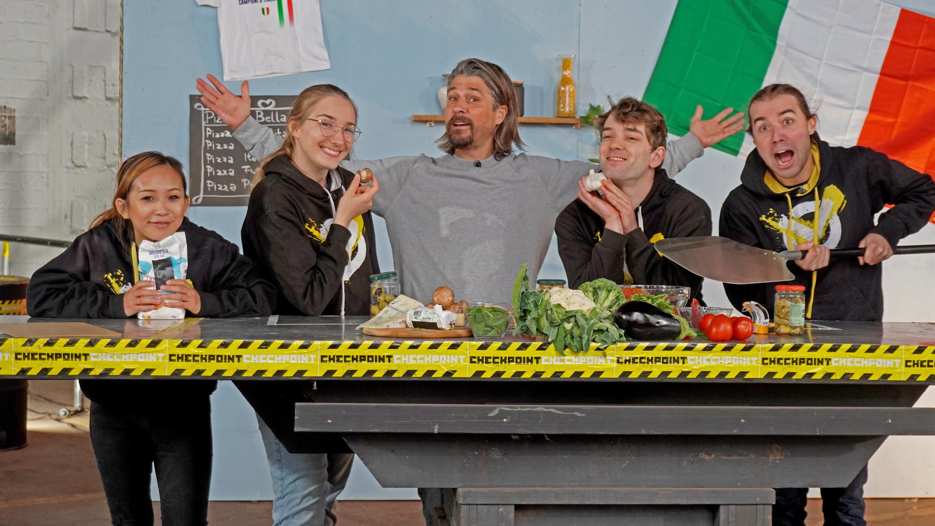 Checkpoint: Das Testteam und Tommy Scheel stehen hinter einem Tisch, auf dem allerlei Zutaten fürs Pizzabacken bereit liegen. 