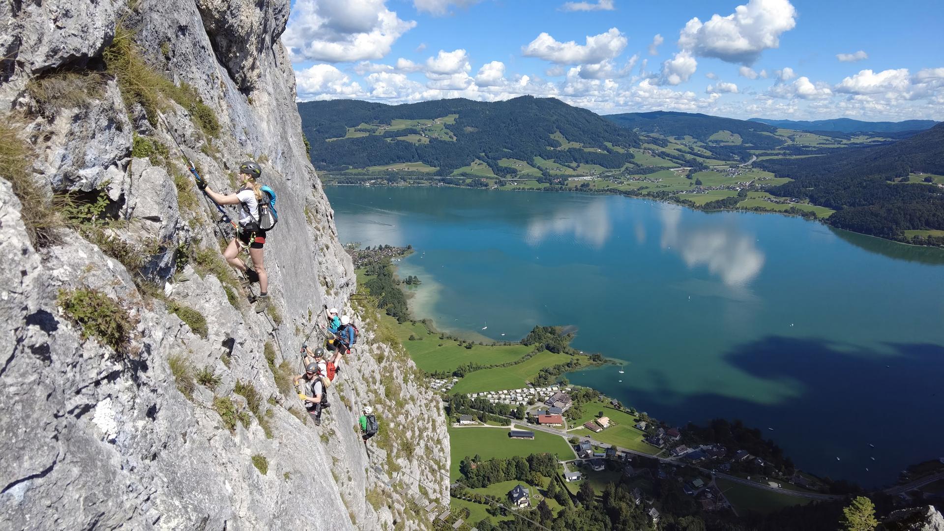 Das Bild zeigt eine Felswand, die Teil des Drachenwand-Klettersteigs in Österreich ist. Auf der linken Seite des Bildes ist eine steile Felsstruktur sichtbar, auf der mehrere Kletterer entlangsteigen. Die Kletterer sind in verschiedenen sportlichen Outfits gekleidet, einige tragen Helme und Klettergurte, während sie sich an einem Drahtseil festhalten.  Im Hintergrund erstreckt sich eine weitläufige Landschaft mit hügeligen Wiesen und einem glitzernden See, der das Licht reflektiert. Der See, möglicherweise der Mondsee, hat eine sanfte bläuliche Farbe und wird von kleinen Booten befahren. Auf der Bodenebene sind einige Gebäude und Campingplätze sichtbar.  Der Himmel ist größtenteils klar mit einigen weißen, flauschigen Wolken. Die gesamte Szene ruft eine Atmosphäre von Abenteuer und Naturverbundenheit hervor. Es ist eine Momentaufnahme von Bergsteigern, die eine beliebte Outdoor-Aktivität ausüben.