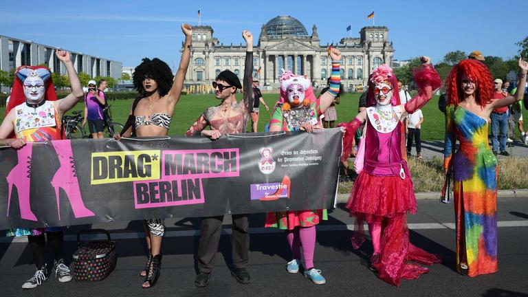 Sechs Drags* stehen vor dem Reichstag in Berlin. Sie halten ein Banner auf dem steht: Drag* March Berlin.