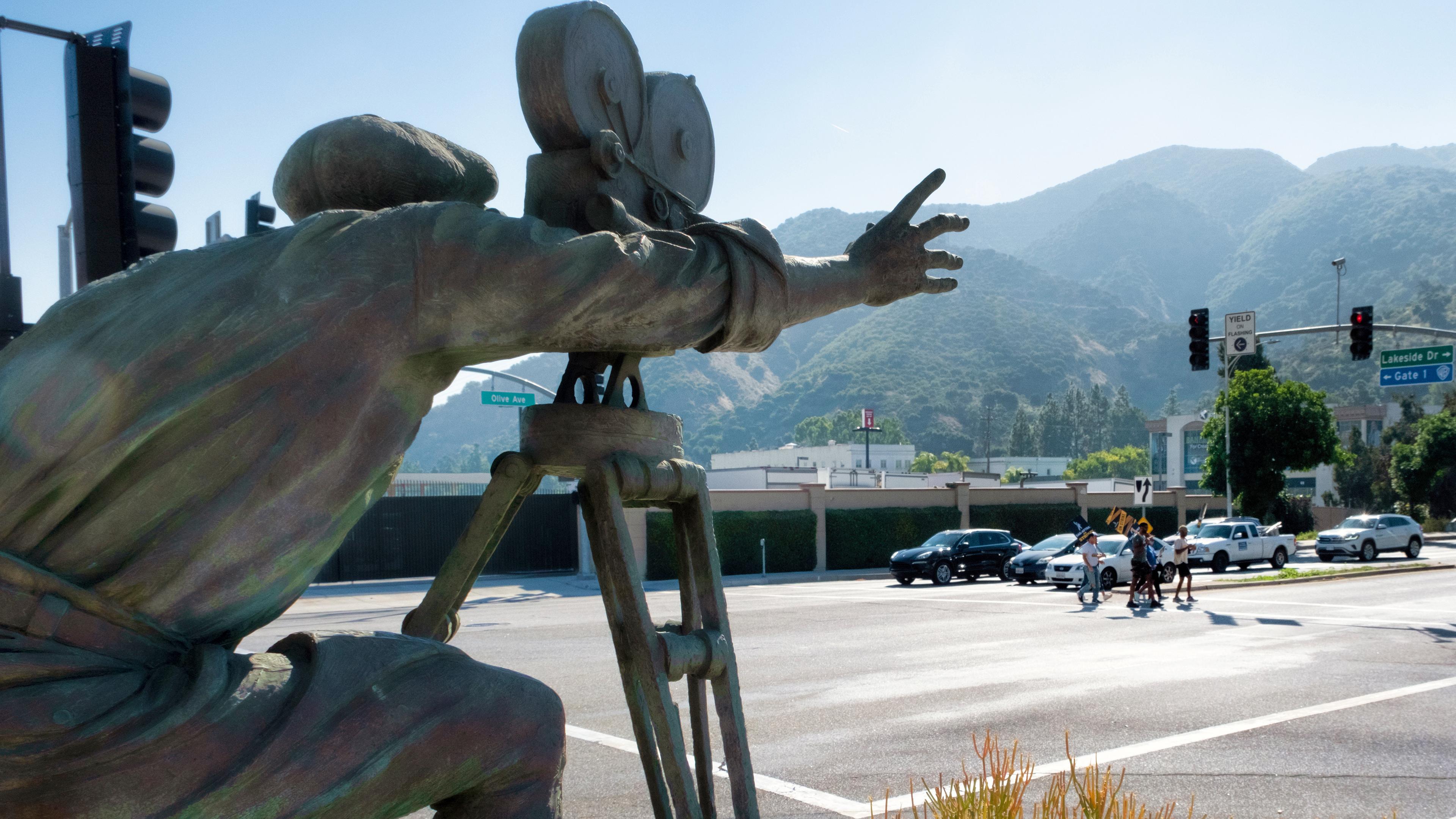 Mit der Statue eines Kameramanns im Vordergrund überqueren SAG-AFTRA-Streikende mit Schildern eine Straße vor den Toren der Warner Bros. Studios, aufgenommen am 26.09.2029