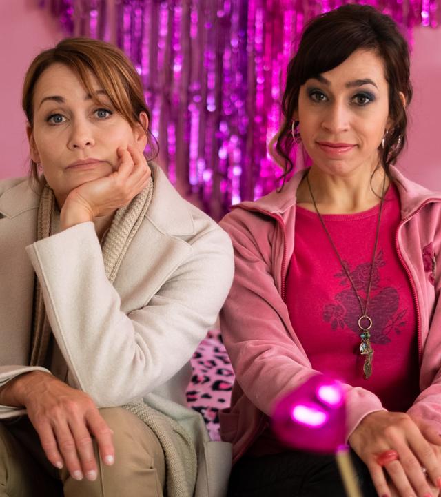 "Dreiraumwohnung": Die beige gekleidete Konstanze (Anja Kling) stützt gefrustet ihren Kopf auf ihre Hand, während sie neben der pink gekleideten, zufrieden lächelnden Jackie (Carol Schuler) auf einem Sofa sitzt. Die Beiden sind von pinkfarbener, glitzernder Wohnungsdekoration umgeben.