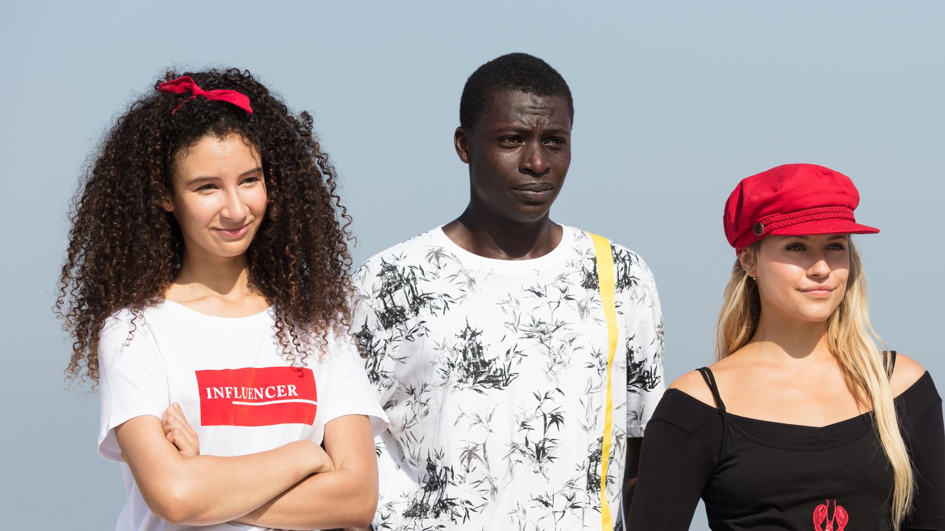 Die Freundesgruppe steht nebeneinander in einer Reihe, die Sonne scheint ihnen ins Gesicht und hinter ihnen ist das Meer zu sehen.