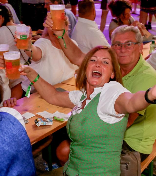 Das Bild zeigt eine lebhafte Szene auf einem Fest in Windischgarsten. Mehrere Personen sitzen an langen Holztischen und halten Becher mit Bier in den Händen. In der Mitte des Bildes ist eine Frau mit braunen Haaren, die in einem traditionellen Dirndlkleid (grünes Mieder und weiße Bluse) gekleidet ist. Sie lächelt breit und hat ihren Arm nach oben gestreckt, als ob sie feiern würde.   Um sie herum sind weitere Menschen sichtbar, einige ebenfalls in Trachten bekleidet. Der Hintergrund ist unklar, aber es gibt eine Atmosphäre von Geselligkeit und Feierlichkeit. Einige Personen heben ihre Becher, während andere in Gespräche vertieft sind. Der Boden scheint ein festlicher Ort zu sein, möglicherweise mit bunten Lichtern und Dekorationen, passend zu einem traditionellen Fest.   Es ist eine fröhliche Umgebung, in der die Leute Spaß haben und die Atmosphäre ausgelassen wirkt.