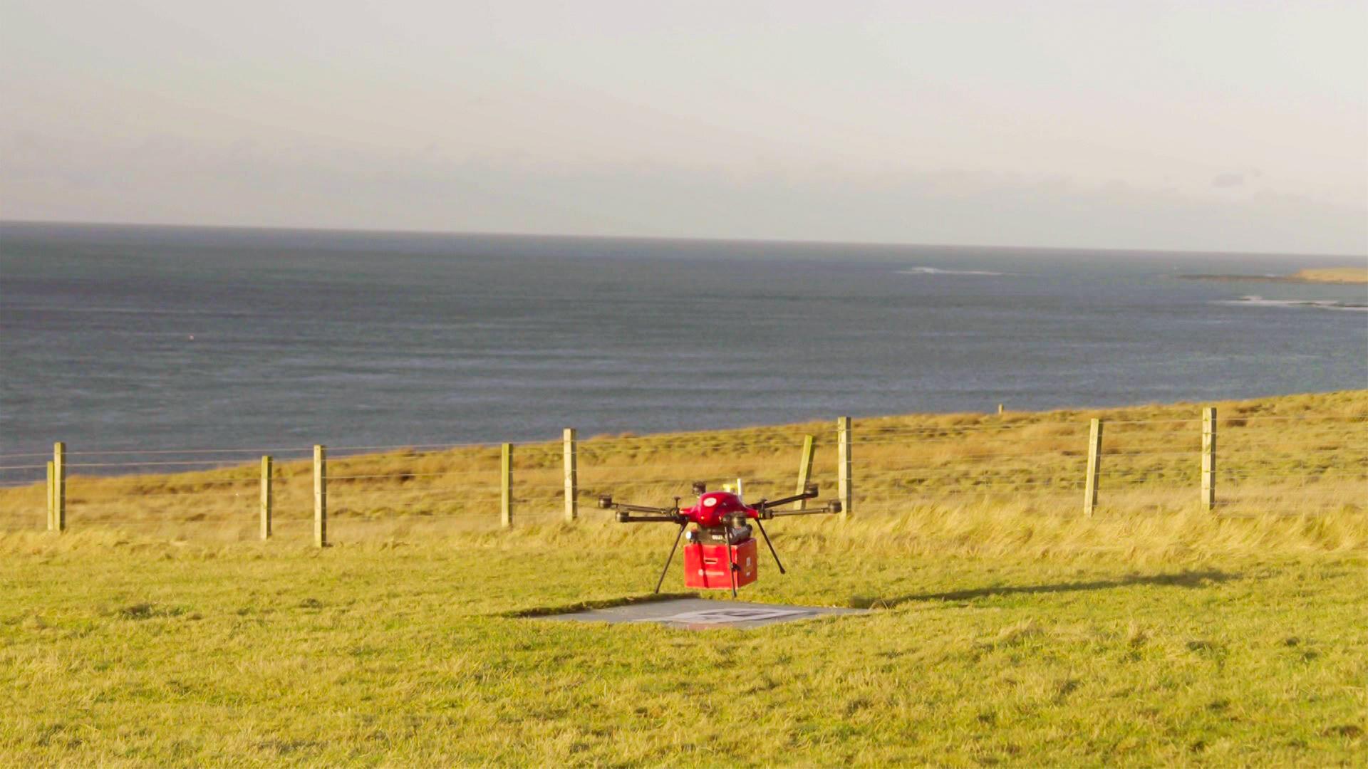 Das Bild zeigt eine leichte, quadratische Drohne, die in einem offenen, grünen Feld auf den schottischen Orkney-Inseln positioniert ist. Die Drohne hat ein rotes Gehäuse und ist über einer markierten Landezone platziert. Im Hintergrund erstreckt sich das Meer, das mit einem sanften Wellengang zu sehen ist. Die Küstenlinie ist am Horizont sichtbar, während sich eine Reihe von Holzpfählen entlang des Feldes zieht, die eine Umzäunung bilden. Der Himmel ist größtenteils klar, mit einigen Wolken, und es scheint Tageslicht zu sein.   Die Szene vermittelt ein Gefühl von Abgeschiedenheit und der technischen Anwendung von Drohnen in ländlichen Gebieten, wo sie zum Transport von wichtigen Gütern eingesetzt werden.