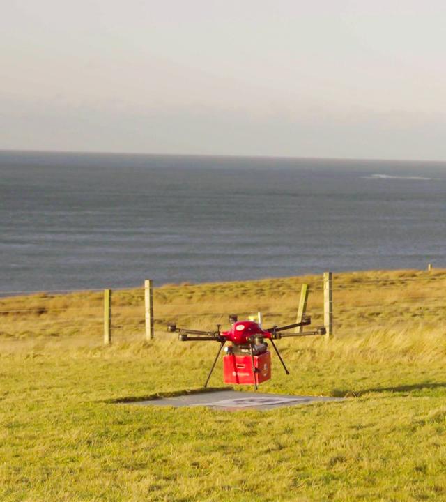 Das Bild zeigt eine leichte, quadratische Drohne, die in einem offenen, grünen Feld auf den schottischen Orkney-Inseln positioniert ist. Die Drohne hat ein rotes Gehäuse und ist über einer markierten Landezone platziert. Im Hintergrund erstreckt sich das Meer, das mit einem sanften Wellengang zu sehen ist. Die Küstenlinie ist am Horizont sichtbar, während sich eine Reihe von Holzpfählen entlang des Feldes zieht, die eine Umzäunung bilden. Der Himmel ist größtenteils klar, mit einigen Wolken, und es scheint Tageslicht zu sein.   Die Szene vermittelt ein Gefühl von Abgeschiedenheit und der technischen Anwendung von Drohnen in ländlichen Gebieten, wo sie zum Transport von wichtigen Gütern eingesetzt werden.