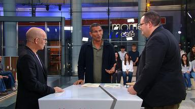 Mitri Sirin steht zwischen Oliver Suchy und Prof. Patrick Glauner beim „Moma duell“. Suchy ist links im Profil zu sehen, Glauner rechts im Vordergrund. Beide sind Sirin zugewandt, der gerade spricht. Im Hintergrund sitzen und stehen Menschen vor einer Glaswand