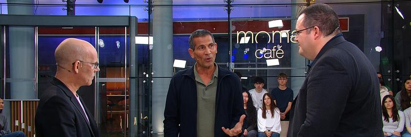 Mitri Sirin steht zwischen Oliver Suchy und Prof. Patrick Glauner beim „Moma duell“. Suchy ist links im Profil zu sehen, Glauner rechts im Vordergrund. Beide sind Sirin zugewandt, der gerade spricht. Im Hintergrund sitzen und stehen Menschen vor einer Glaswand