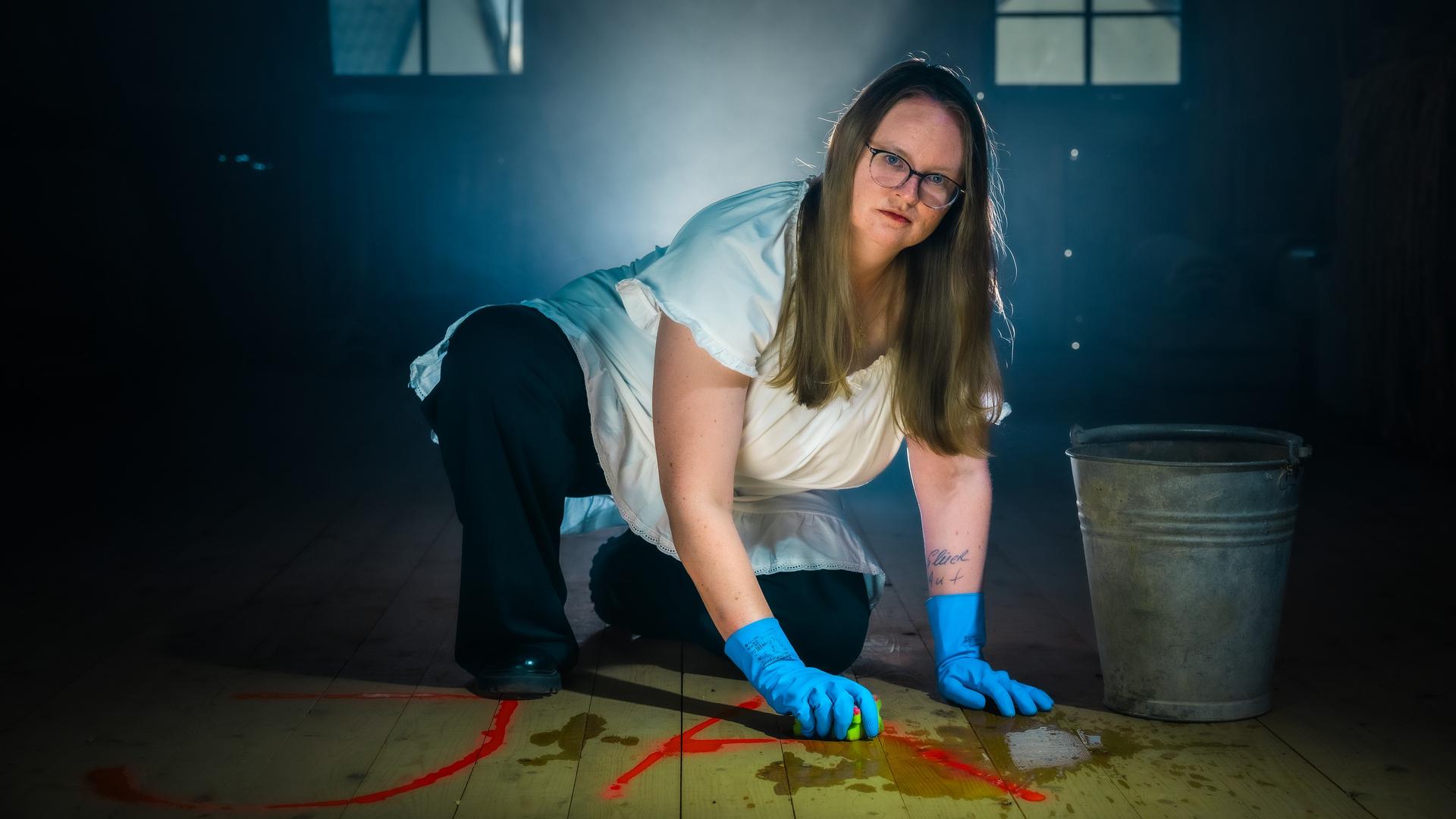 Laura kniet auf dem Boden und putzt mit Handschuhen ein Ja vom Boden. Dabei schaut sie ernst in die Kamera.