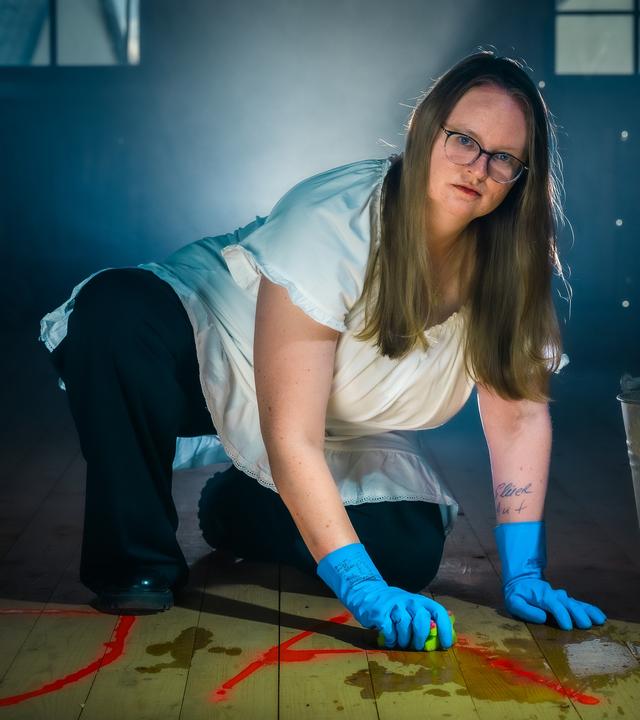 Laura kniet auf dem Boden und putzt mit Handschuhen ein Ja vom Boden. Dabei schaut sie ernst in die Kamera.