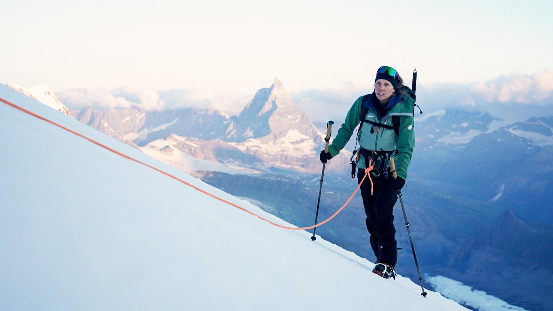 Eine Bergsteigerin mit grüner Jacke und Rucksack steigt mit Skistöcken und Seilsicherung eine schneebedeckte Steigung hinauf. Im Hintergrund sind das Matterhorn und weitere schneebedeckte Berge sichtbar.  