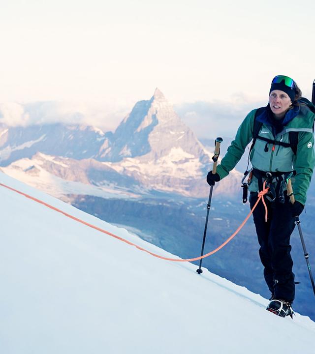 Eine Bergsteigerin mit grüner Jacke und Rucksack steigt mit Skistöcken und Seilsicherung eine schneebedeckte Steigung hinauf. Im Hintergrund sind das Matterhorn und weitere schneebedeckte Berge sichtbar.  