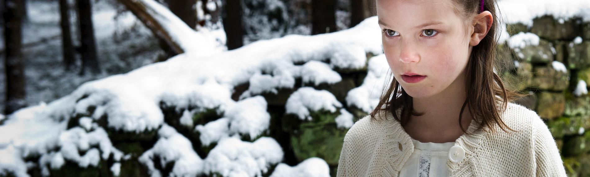 Hanna (Greta Oceana Dethlefs) als Kind steht vor einer verschneiten Steinmauer. Dahinter sind Bäume zu sehen.