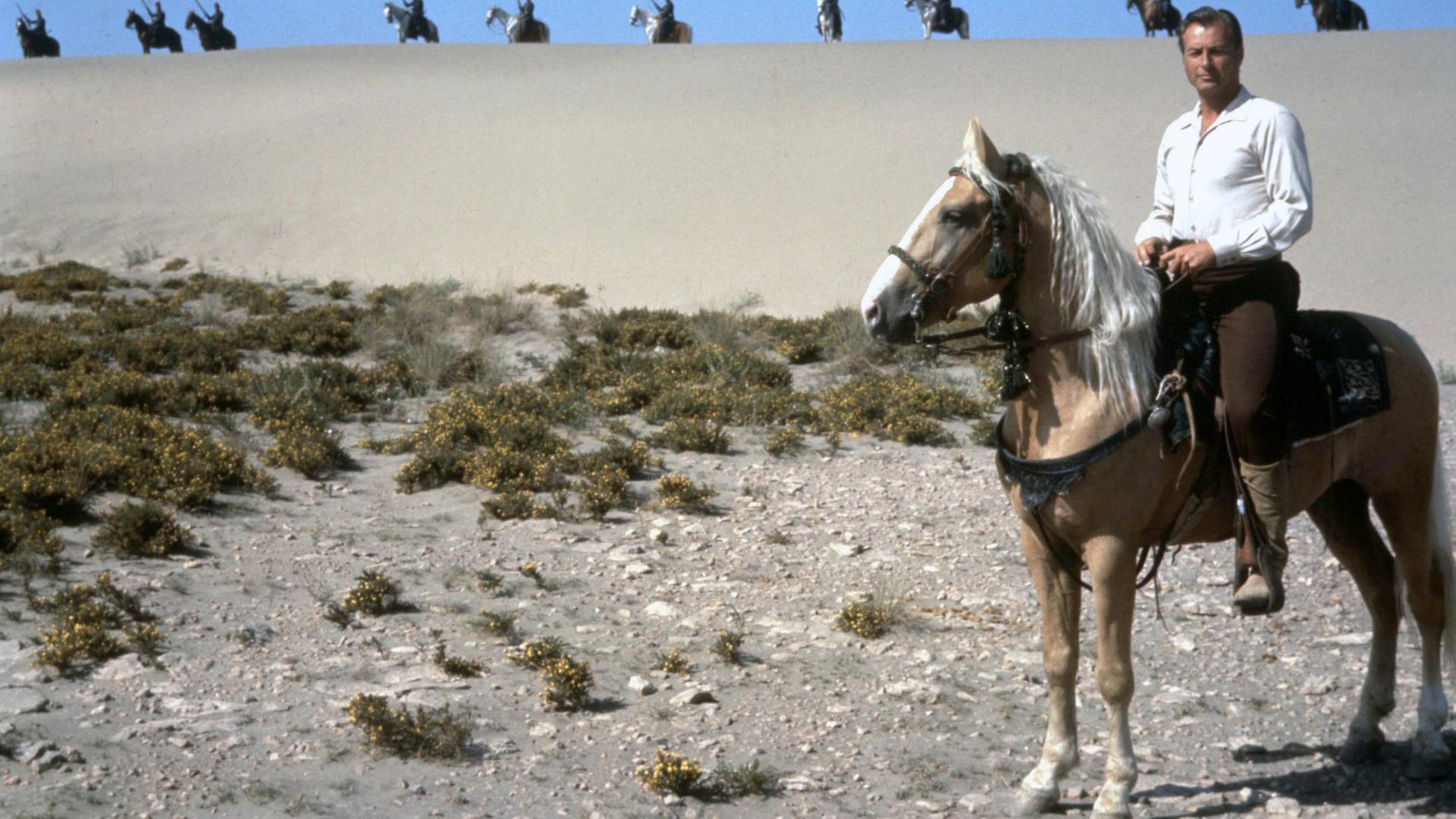 Kara Ben Nemsi (Lex Barker) sitzt auf einem Pferd und schaut in die Ferne. Hinter ihm auf einer Sanddüne sieht man mehrere bewaffnete Krieger, ebenfalls auf Pferden.
