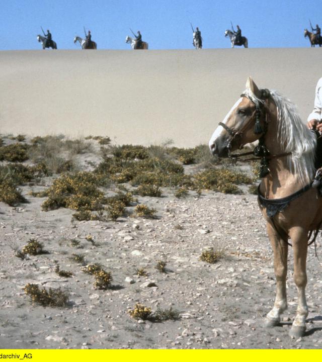 Kara Ben Nemsi (Lex Barker) sitzt auf einem Pferd und schaut in die Ferne. Hinter ihm auf einer Sanddüne sieht man mehrere bewaffnete Krieger, ebenfalls auf Pferden.