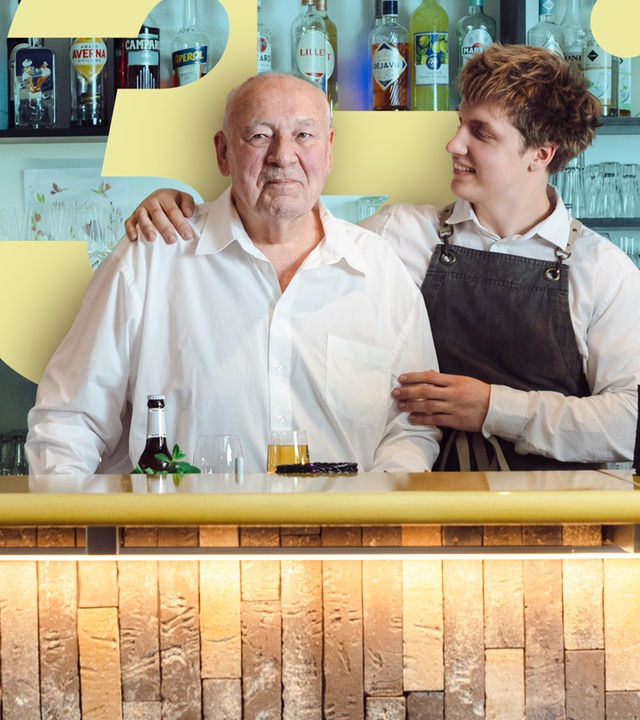 Ein alter Mann hinter einem Bar-Tresen, neben ihm ein jüngerer Barmann, der ihn lächelnd anschaut und ihm die Hand auf die Schulter legt. 