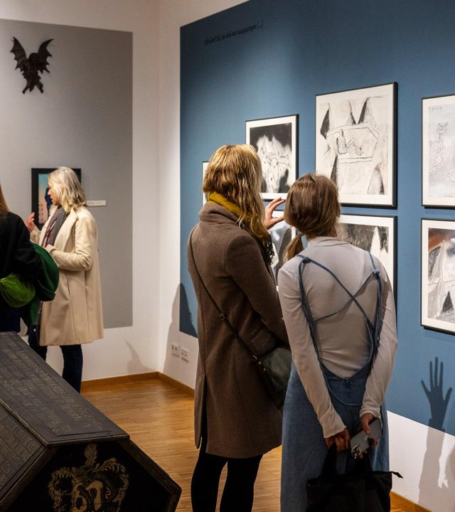 In einem Ausstellungsraum betrachten vier Frauen in Zweiergrüppchen Bilder an den Wänden. In der Mitte des Raums steht ein Holzsarg.