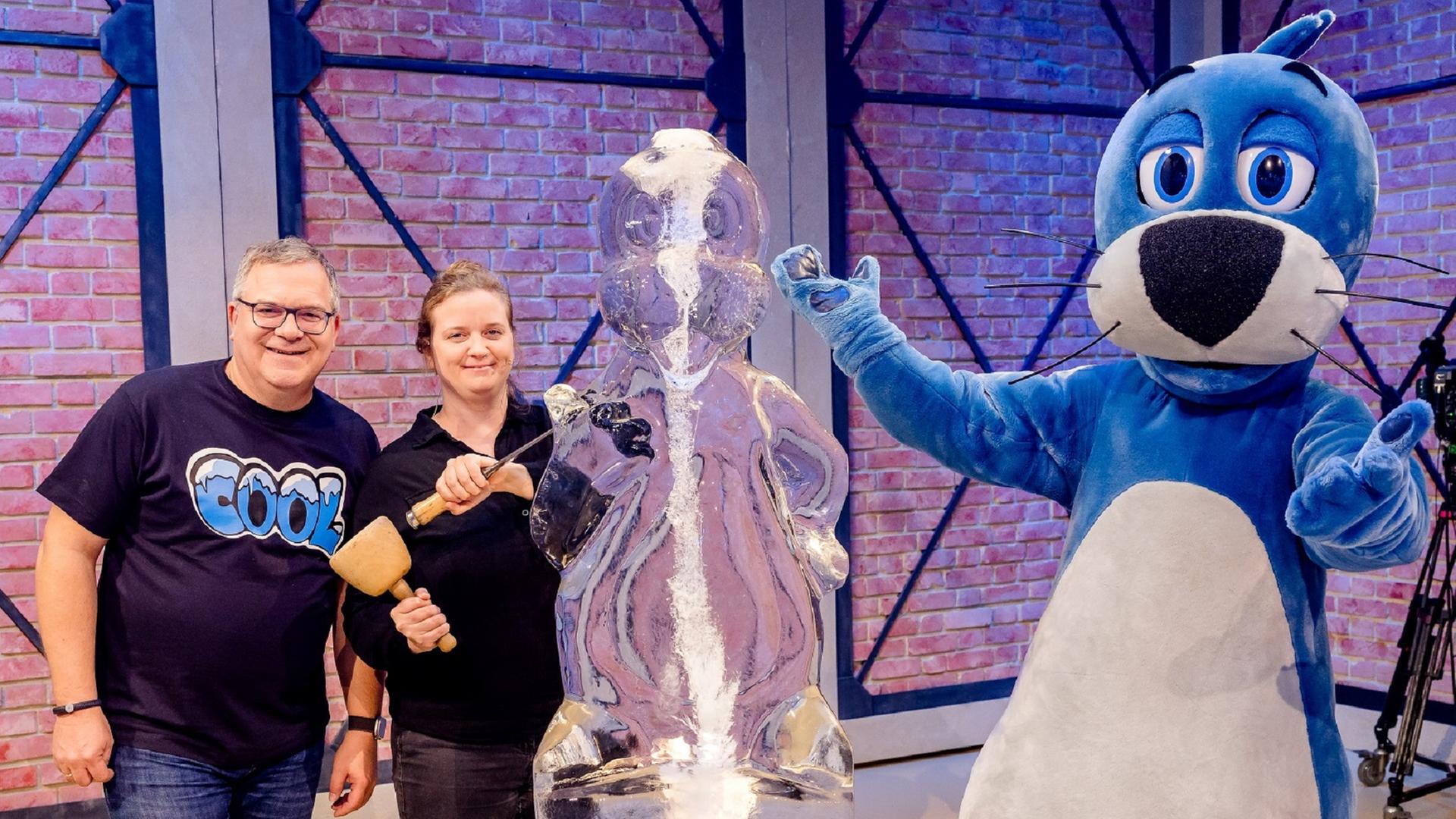 Die Eisbildhauerin Lea Diehl (m.) steht mit Elton (l.) und Piet Flosse (r.) vor einer Piet Flosse-Figur aus Eis. In den Händen hält sie einen Meißel und einen Holzhammer.